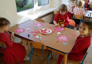 Widok na siedzące przy dwóch stolikach dzieci, które wyklejają różowe koła czerwonymi serduszkami.